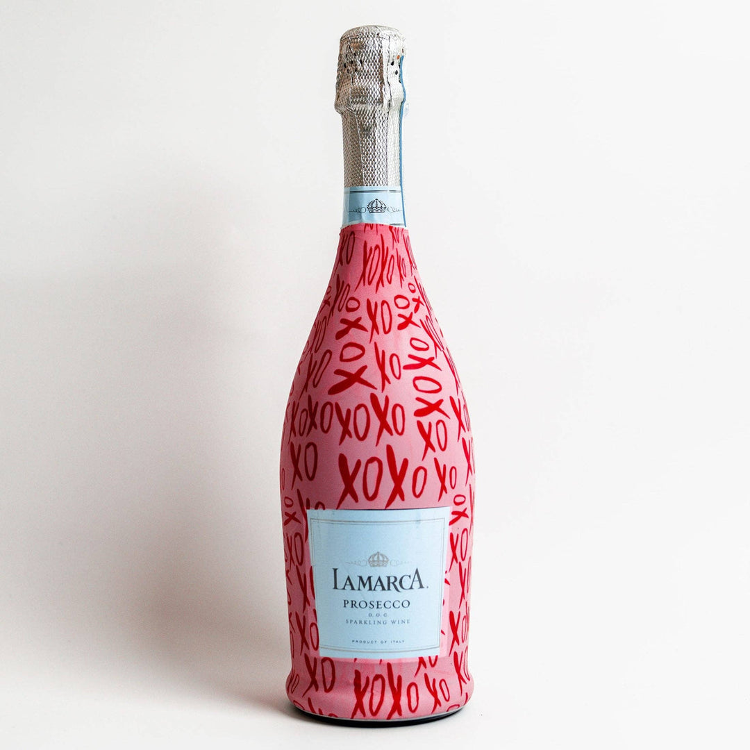 Hand-painted glass bottle of La Marca Prosecco in a studio setting featuring a bold red script XOXO pattern on a bubblegum pink wrap with a signature light blue label and textured silver foil neck
