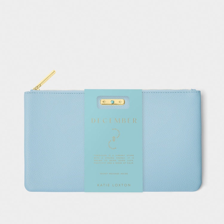 Pebbled vegan leather pouch by Katie Loxton featuring a sky blue December birthstone clutch centered on a neutral backdrop with a small turquoise stone set in a gold bar, a gold-tone zipper, and a branded teal paper wrap with gold foil lettering