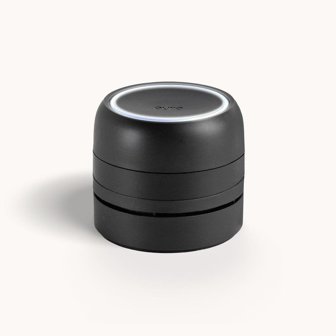 Product photograph of the Pura smart fragrance diffuser featuring a matte black tiered cylindrical design, a glowing white LED circular ring on top, and the brand name subtly embossed on the surface against a white background