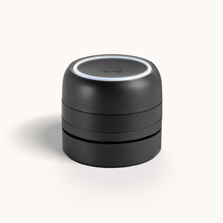 Product photograph of the Pura smart fragrance diffuser featuring a matte black tiered cylindrical design, a glowing white LED circular ring on top, and the brand name subtly embossed on the surface against a white background