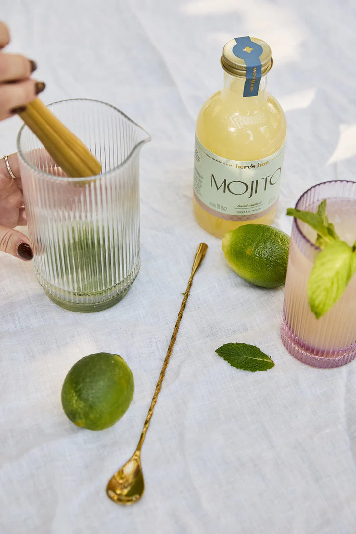 Lifestyle photograph of mojito cocktail preparation on a white linen outdoor surface featuring a fluted glass pitcher, a long gold twisted bar spoon, and fresh green limes scattered near a bottle of Here&