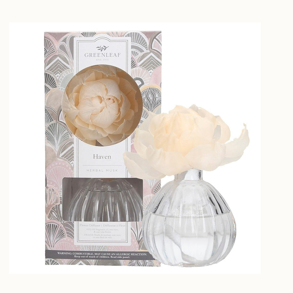 Studio product photograph by Greenleaf displaying the Haven Herbal Musk flower diffuser featuring a large cream-colored sola flower resting on a clear bulbous ribbed glass vase beside a decorative gift box with a pink and grey art deco fan pattern