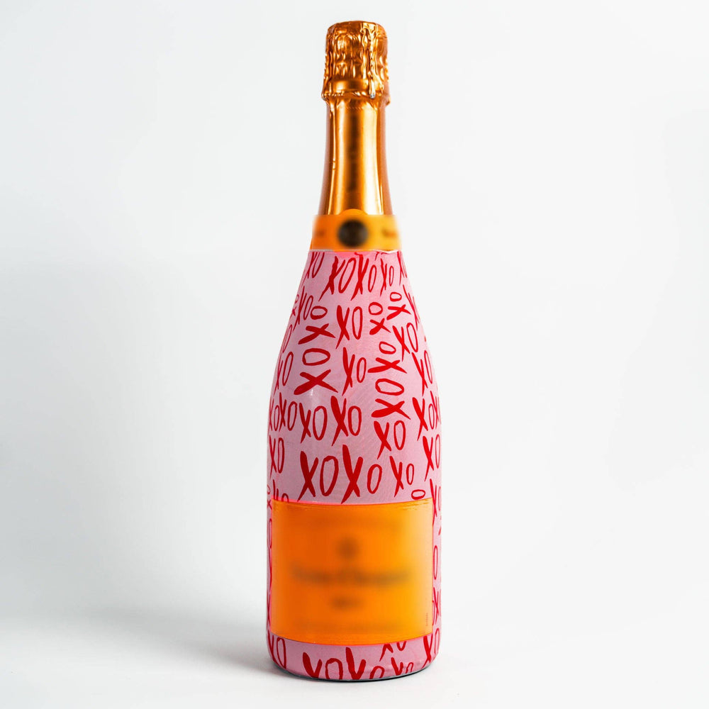 Studio photograph of a Veuve Clicquot champagne bottle featuring a pink wrap with red XOXO calligraphy, iconic orange branding, and a shimmering gold foil capsule against a clean white background