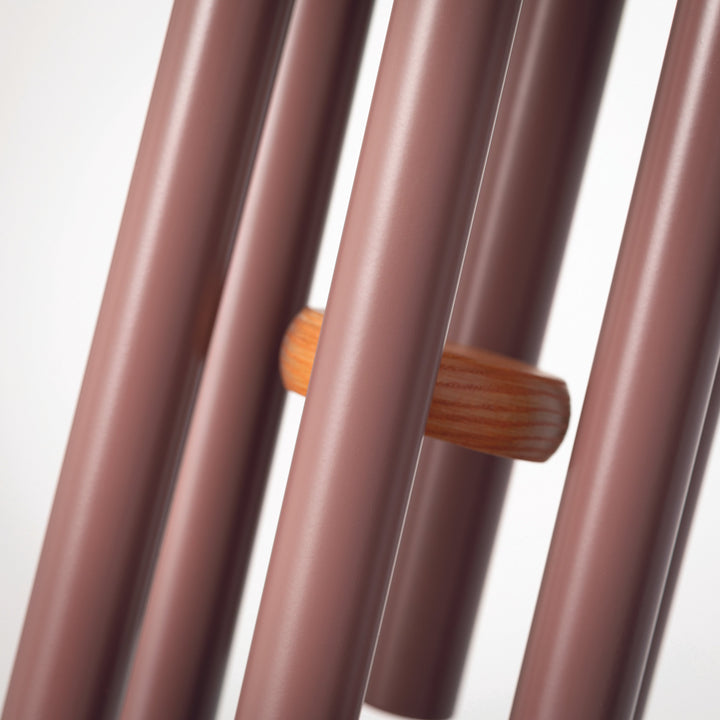 Photograph of metal and wood wind chimes featuring matte plum-colored tubes and a central orange-toned wooden striker with visible grain patterns against a minimalist white studio background