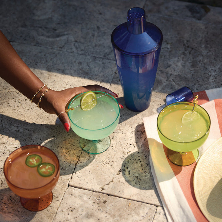 A lifestyle photograph captures vibrant margaritas and a cobalt blue shaker on a sunlit stone patio featuring a hand with red nails holding a teal glass and an amber drink garnished with fresh jalapeño slices