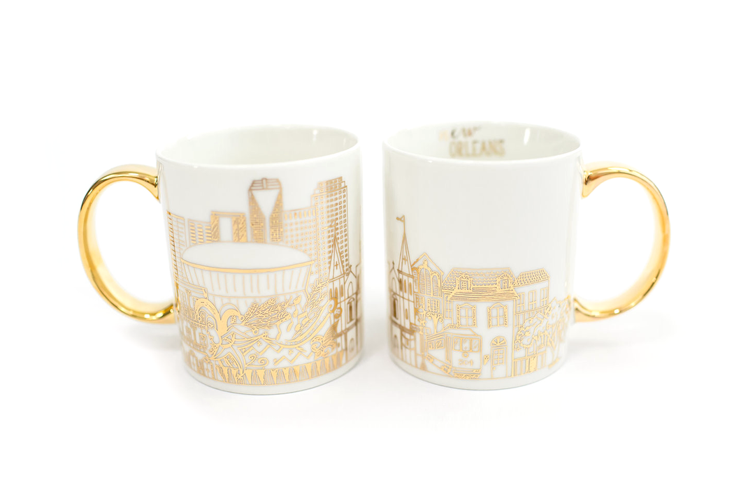 White ceramic mugs featuring gold metallic line art of New Orleans landmarks including the Caesars Superdome and St. Louis Cathedral with polished gold handles and the city name printed on the inner rim