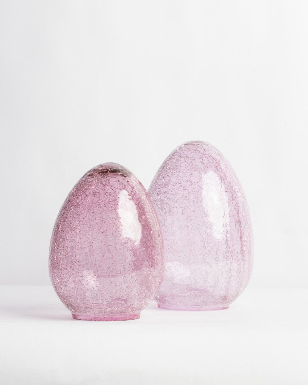 Hand-blown crackle glass sculptures of two translucent pink egg-shaped vessels positioned on a minimalist white studio surface featuring intricate internal webbing and glossy light reflections