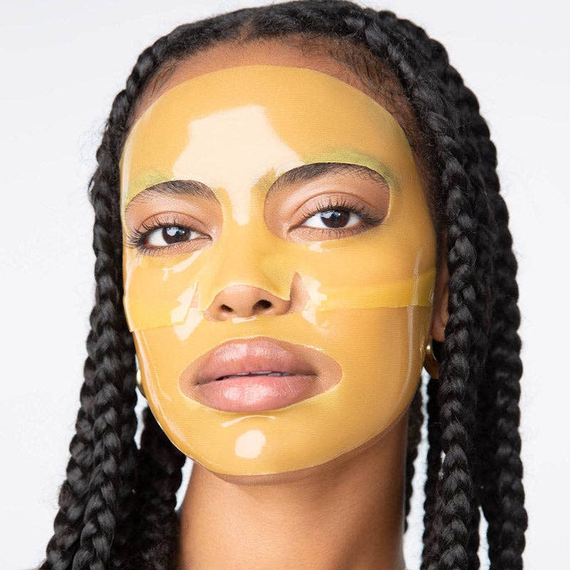 Digital photograph of a Black woman with long dark braids in a studio wearing a translucent yellow hydrogel face mask with a glossy golden sheen and precision cutouts around her eyes and mouth