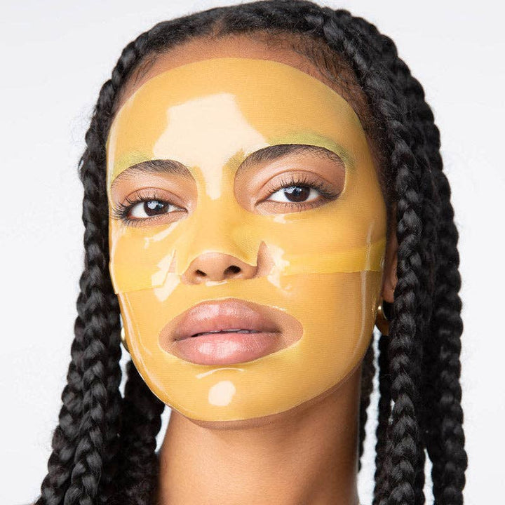 Digital photograph of a Black woman with long dark braids in a studio wearing a translucent yellow hydrogel face mask with a glossy golden sheen and precision cutouts around her eyes and mouth