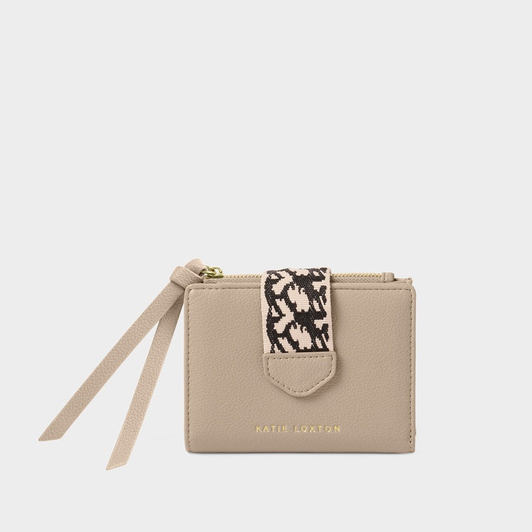 Product photography of a Katie Loxton vegan leather wallet in taupe on a neutral studio background, featuring a monochrome geometric woven strap, gold-toned hardware, and double long tassel zip pulls