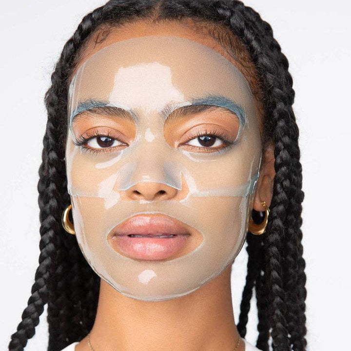 Digital photograph of a woman with long black braids wearing a transparent hydrogel face mask in a studio setting, featuring gold hoop earrings and glossy pink lips