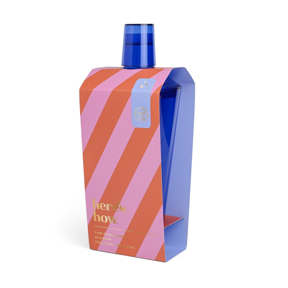Product photograph of The Penelope Riviera Cocktail Shaker by Here's How featuring a translucent cobalt blue vessel within a pink and orange diagonal striped sleeve with gold foil lettering against a white studio background