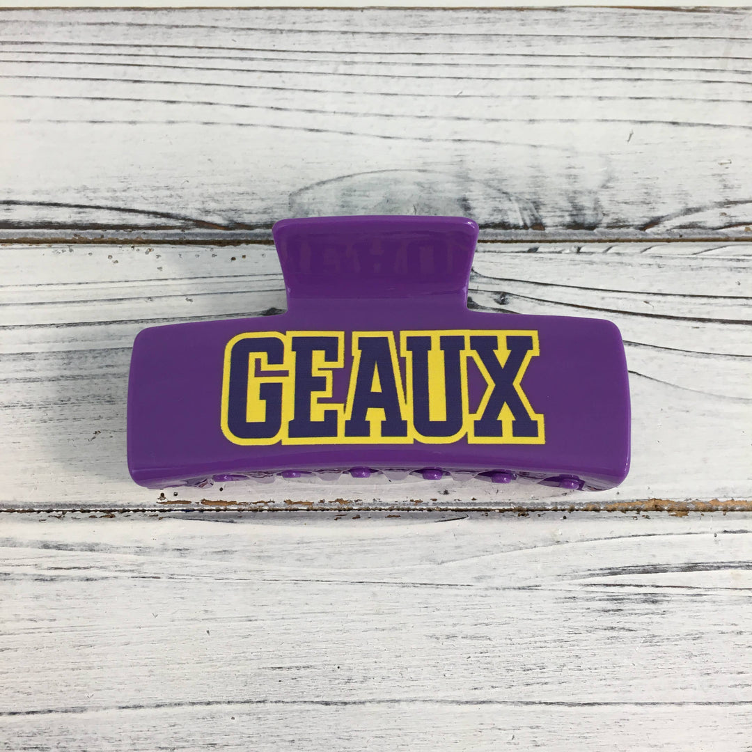 Plastic hair claw clip on a white wooden background featuring yellow GEAUX block lettering with a purple outline and a glossy finish