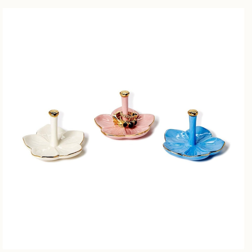 Ceramic flower-shaped ring holders in cream, pink, and blue arranged on a white background featuring gold-trimmed petals, vertical storage pillars, and a cluster of gold rings on the center pink dish