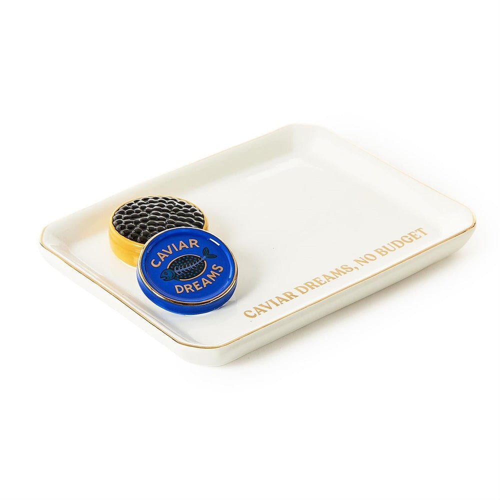Porcelain trinket tray by Jonathan Adler featuring a sculptural miniature caviar tin with black beads and a blue lid next to gold-foil lettering that says CAVIAR DREAMS NO BUDGET on a white background with a metallic gold rim