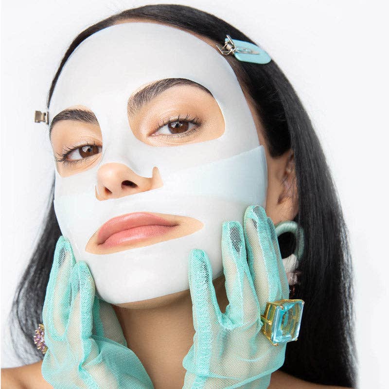 Digital photograph of Camila Mendes wearing a white facial sheet mask in a studio setting featuring sheer mint green mesh gloves and a large square-cut aquamarine ring