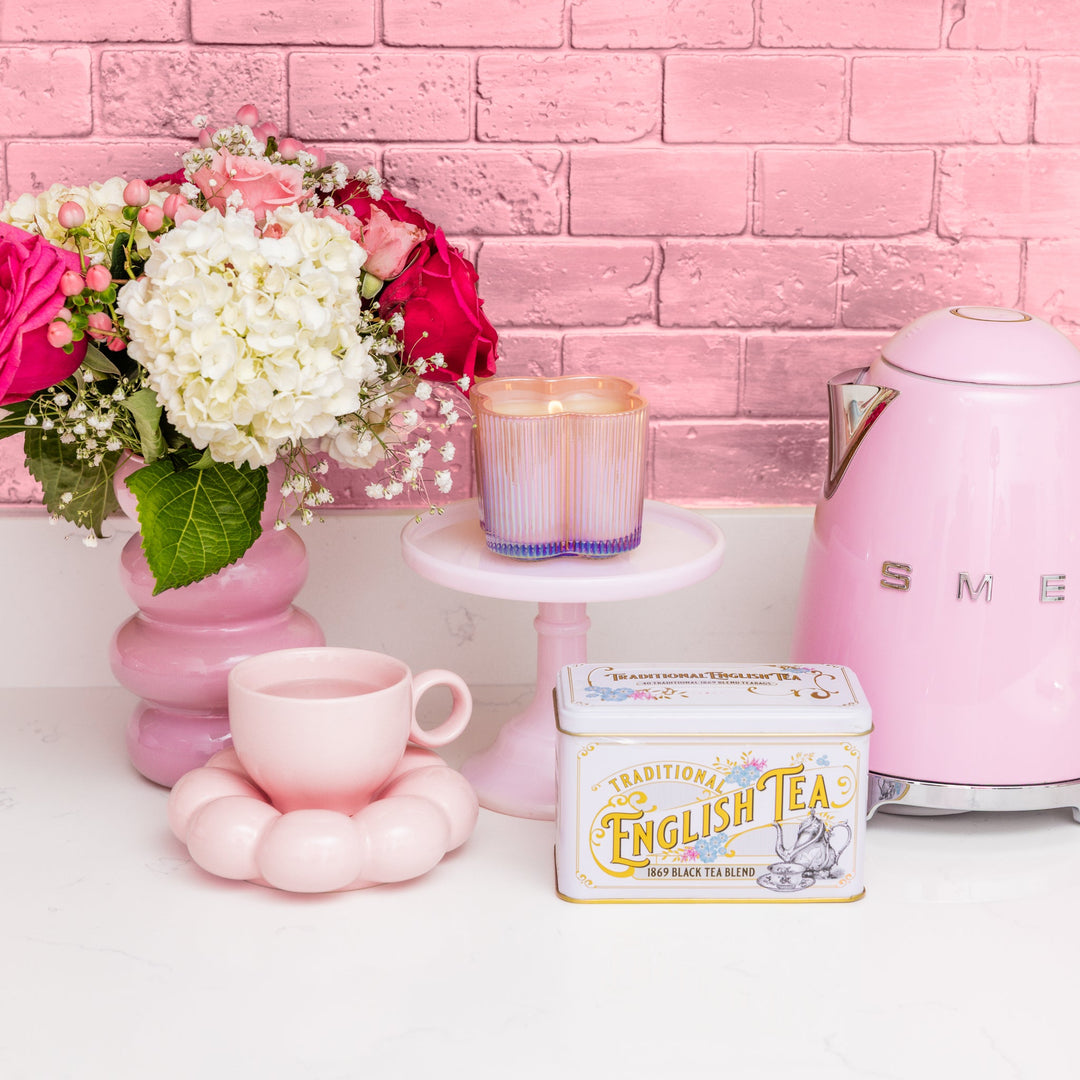 Digital photograph of a pink-themed tea arrangement on a white kitchen counter featuring a pink Smeg electric kettle, a Traditional English Tea tin, and a whimsical cloud-shaped teacup set against a metallic pink brick wall