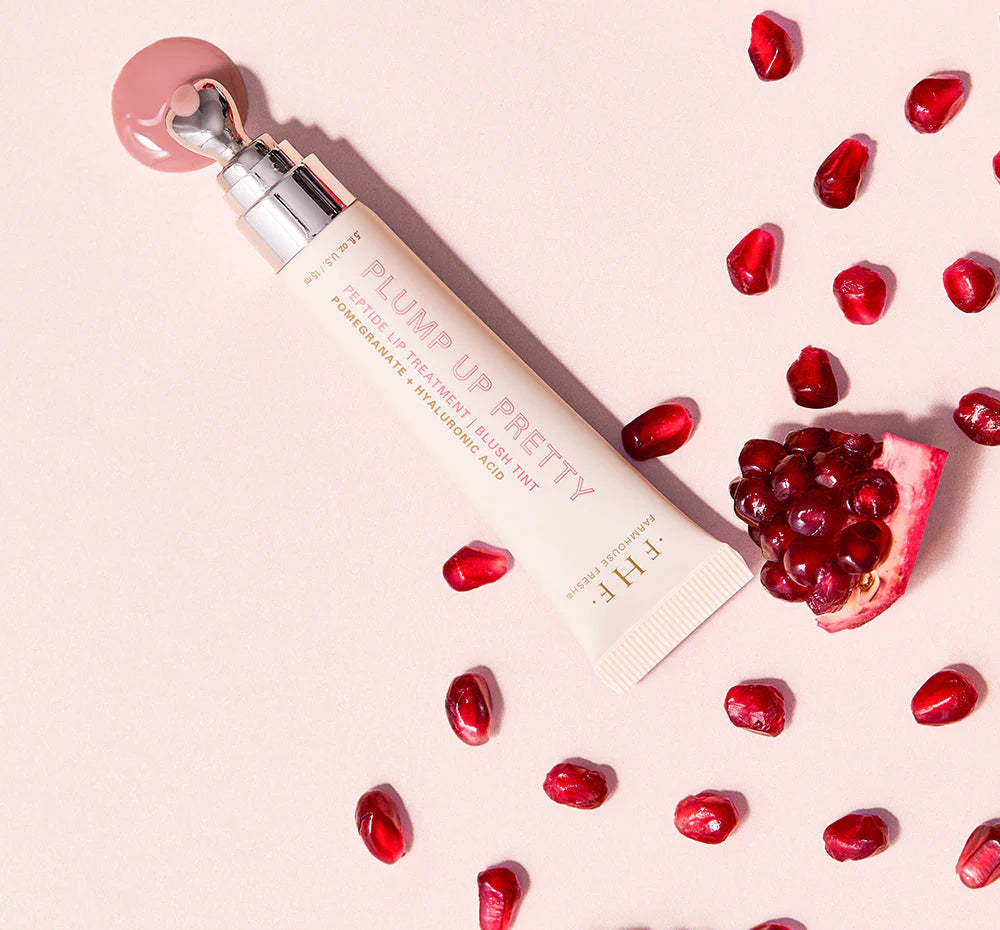 Product photograph by Farmhouse Fresh of a Plump Up Pretty Peptide Lip Treatment tube on a soft pink background featuring a metallic applicator tip resting in pink gloss surrounded by scattered ruby-red pomegranate seeds and a sliced fruit wedge