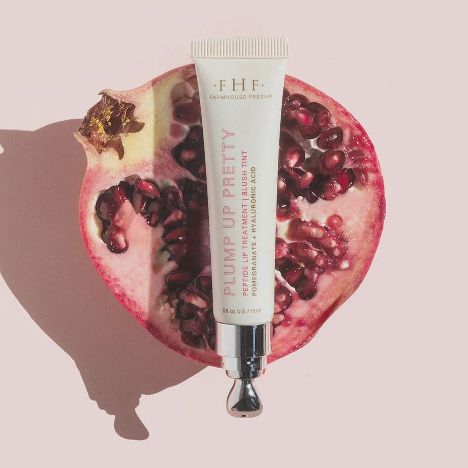 Studio photography by Farmhouse Fresh showcasing the Plump Up Pretty Peptide Lip Treatment tube centered on a halved pomegranate filled with vibrant red seeds and featuring a sleek silver applicator tip against a pastel pink backdrop