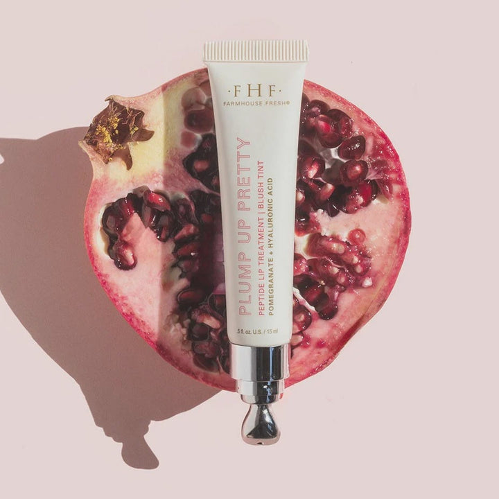 Studio photography by Farmhouse Fresh showcasing the Plump Up Pretty Peptide Lip Treatment tube centered on a halved pomegranate filled with vibrant red seeds and featuring a sleek silver applicator tip against a pastel pink backdrop