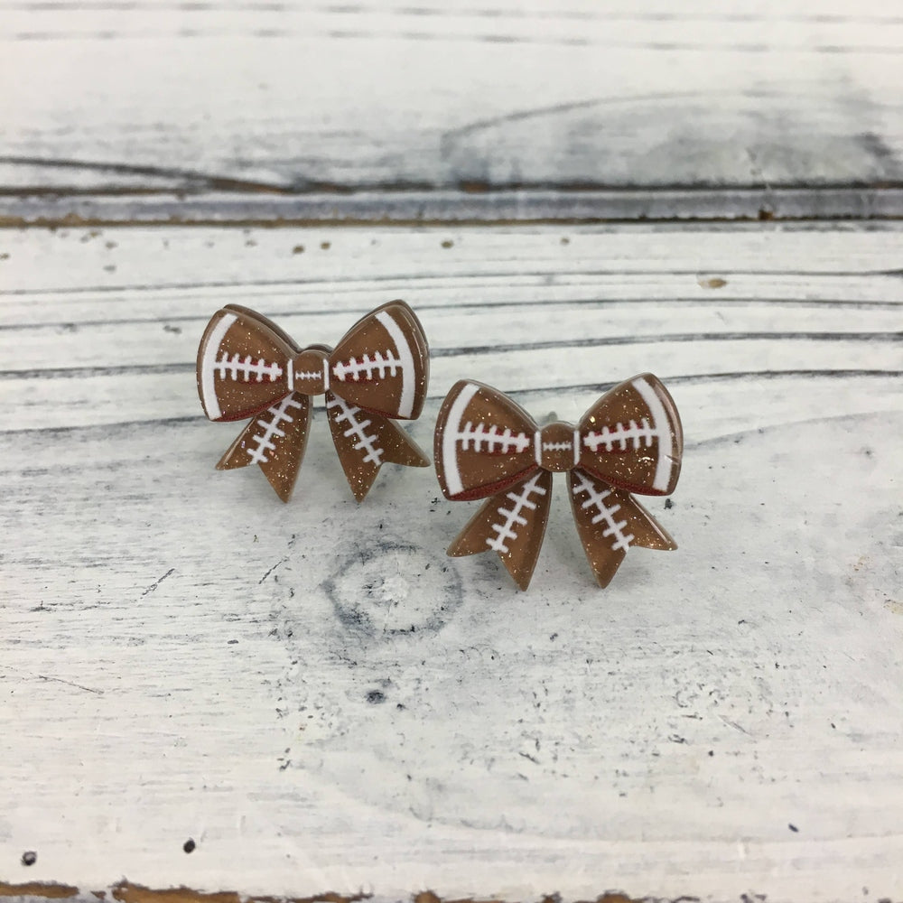 Acrylic resin stud earrings featuring brown football-themed bows with white laces and gold glitter accents are displayed on a rustic white wooden tabletop