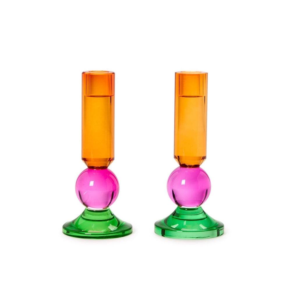 Hand-blown borosilicate glass candle holders by Maison Balzac featuring a pair of vibrant pedestals against a white background with amber cylindrical tops, translucent magenta pink spheres, and emerald green flared bases