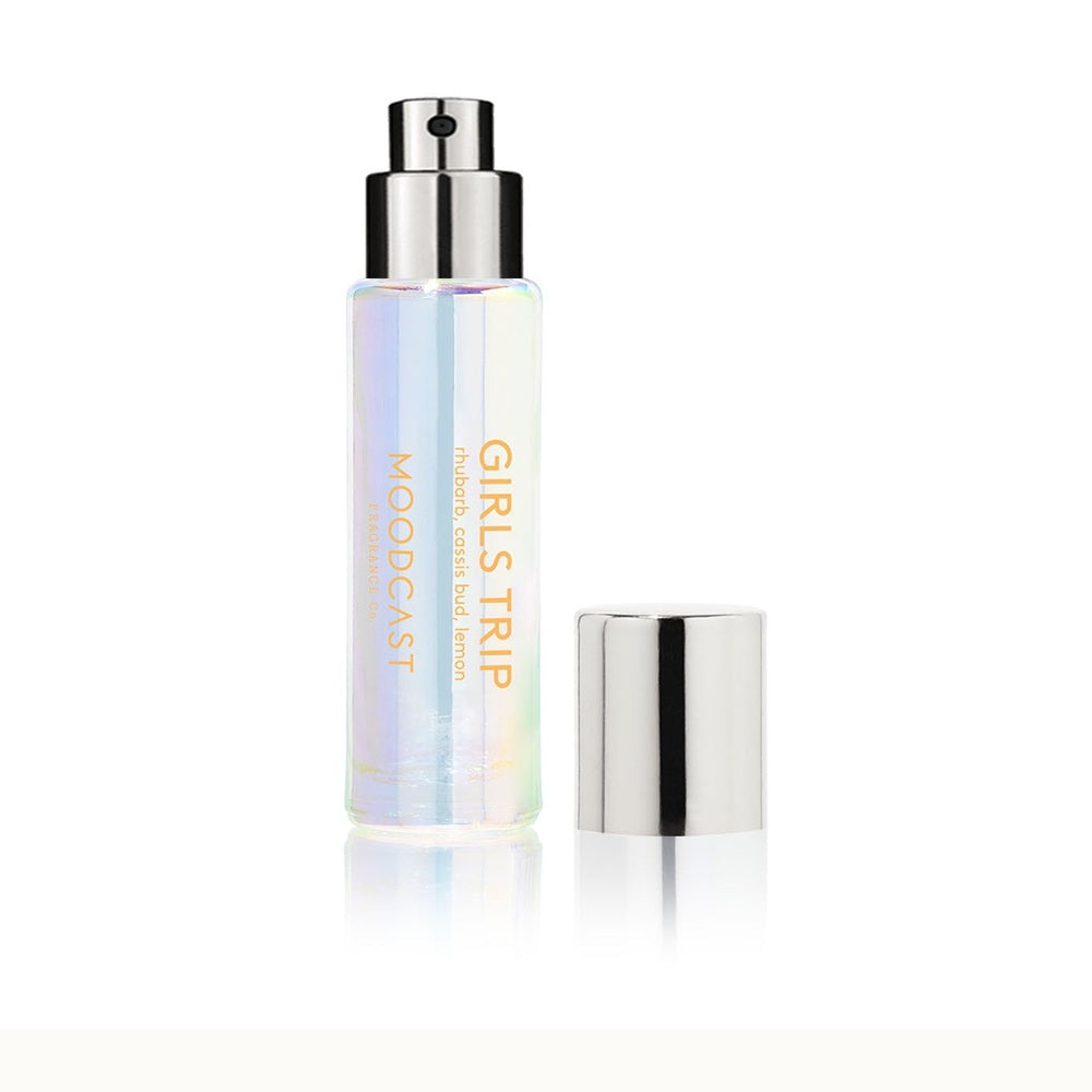 Product photograph by Moodcast Fragrance Co of an iridescent Girls Trip perfume bottle on a reflective white surface featuring a silver spray nozzle, separate metallic cap, and orange text listing rhubarb, cassis bud, and lemon notes