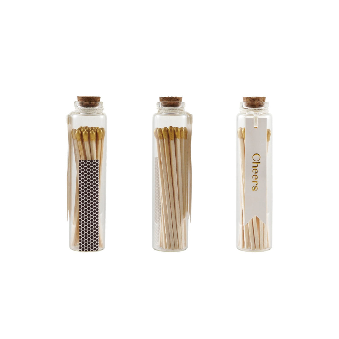 Product photograph of three glass vials filled with gold-tipped matches against a white background, featuring natural cork stoppers, a honeycomb strike strip, and a white tag with gold Cheers text