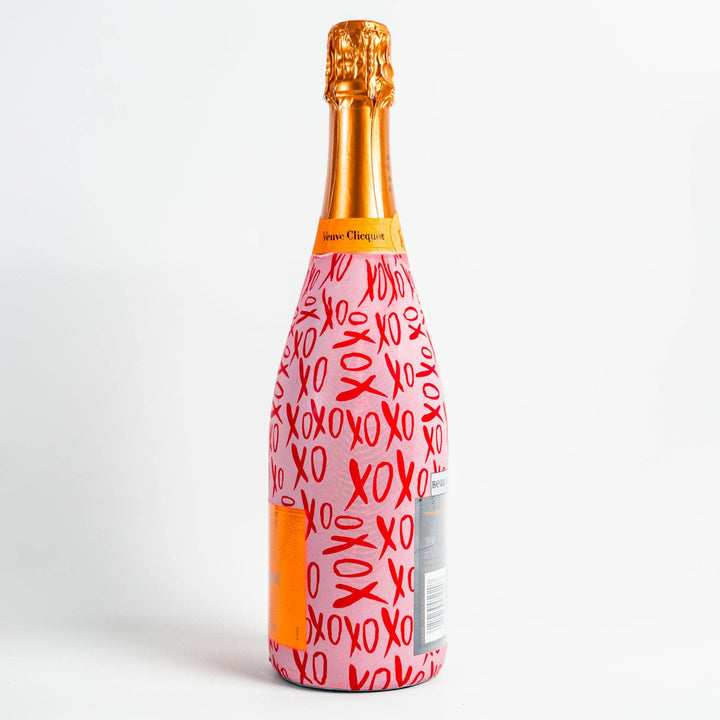 Photograph of a Veuve Clicquot champagne bottle on a white background featuring a pink decorative sleeve with red XOXO script, a signature orange label, and gold foil wrapping the cork