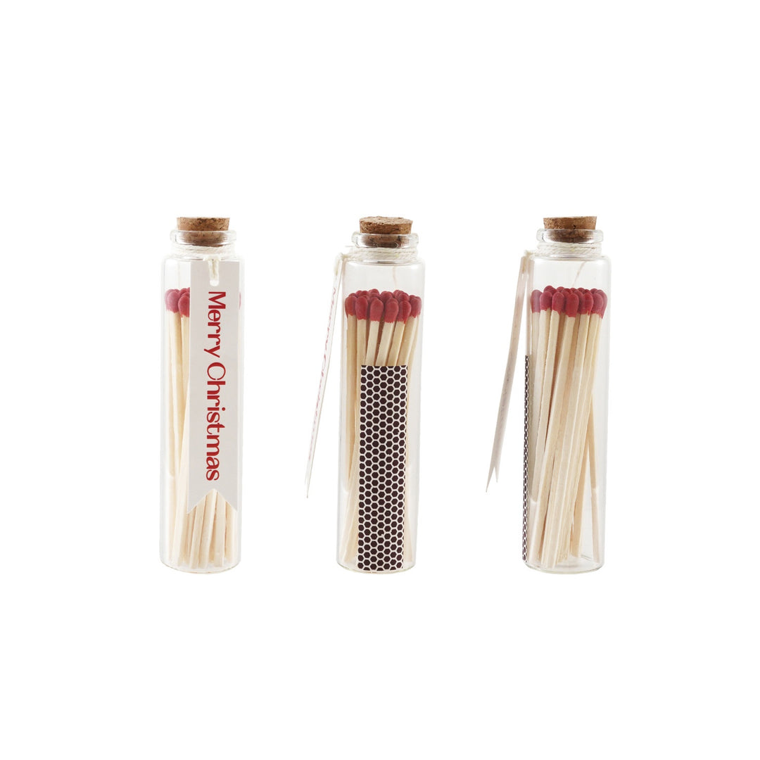 Studio photograph of three corked glass apothecary jars filled with red-tipped matches featuring a white Merry Christmas tag and a black honeycomb striking surface on a white background