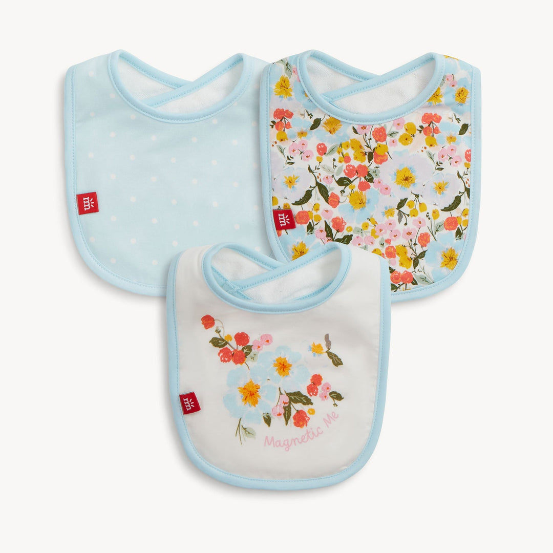 Fabric baby bibs by Magnetic Me displayed on a white background featuring light blue polka dots, a dense multicolored wildflower pattern, and a central floral bouquet with pink script text and signature red brand tags