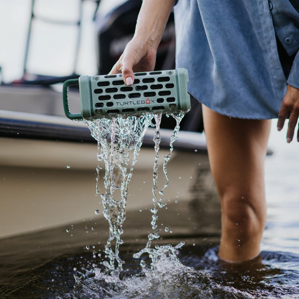 Digital lifestyle photograph of a sage green Turtlebox speaker held by a person wearing a light blue corduroy shirt standing in water next to a boat with water pouring out of the speaker’s honeycomb grille