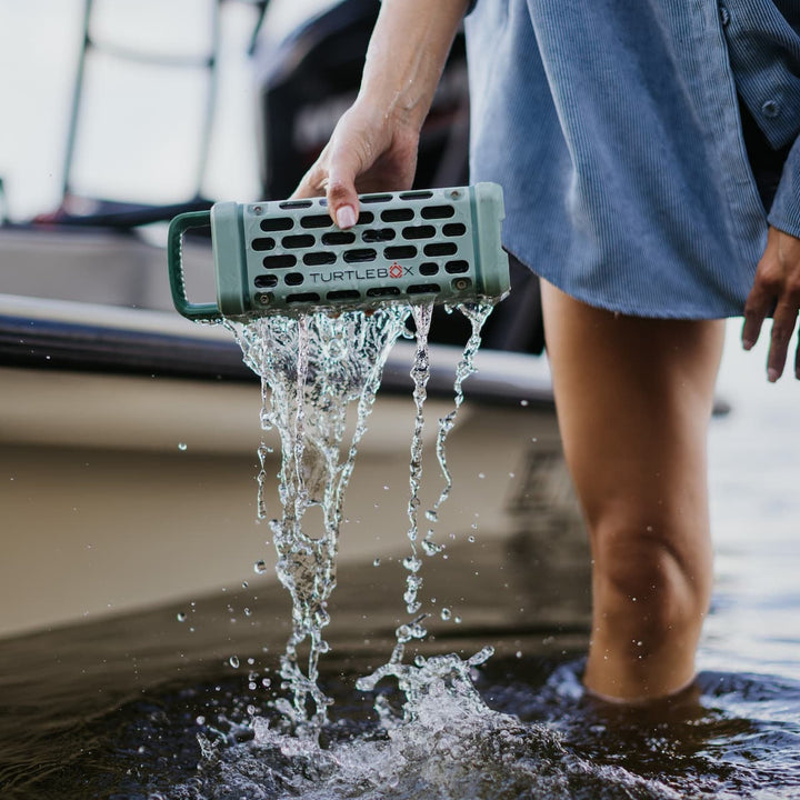 Digital lifestyle photograph of a sage green Turtlebox speaker held by a person wearing a light blue corduroy shirt standing in water next to a boat with water pouring out of the speaker’s honeycomb grille