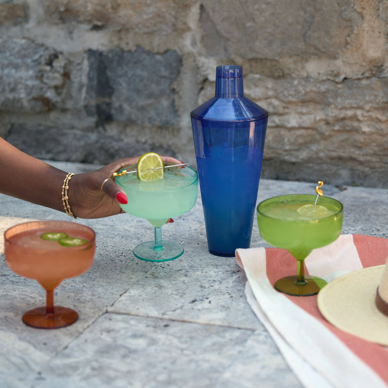 Photograph of vibrant summer cocktails and a blue translucent shaker on a stone patio featuring a hand with red nails holding a lime-garnished margarita, a pink drink with sliced jalapeños, and a green cocktail accented by a golden flamingo stirrer
