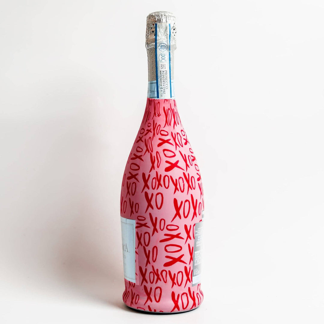 Graphic-designed pink Prosecco bottle art positioned in a studio setting featuring a repeating red XOXO graffiti pattern, a textured silver foil cap, and a vertical light blue DOC seal