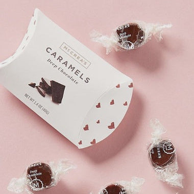 Commercial product photograph by McCrea's Candies of Deep Chocolate Caramels on a pink background featuring a white pillow box with red heart patterns and scattered individually twist-wrapped candies