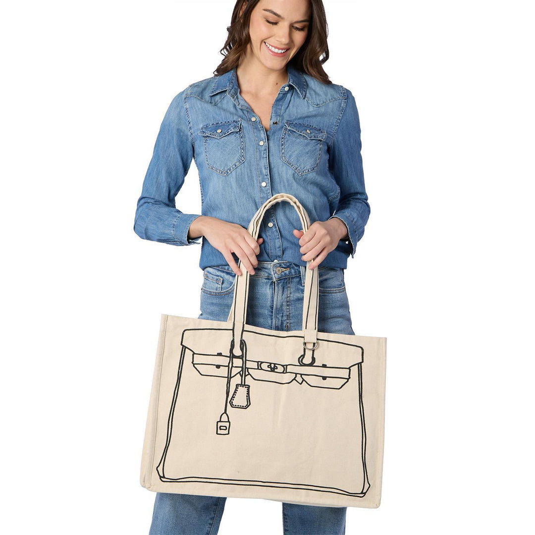 Photograph of a smiling woman in a studio holding a large cream canvas tote bag featuring a black line-drawn illustration of a luxury Birkin-style handbag with a detailed lock and hanging key fob tag