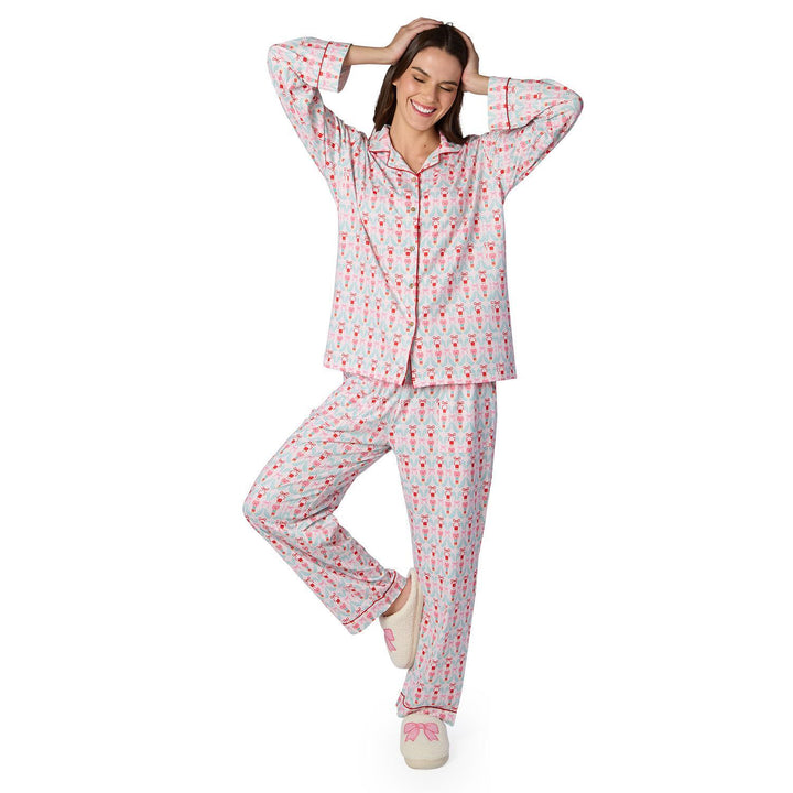 Photograph of a smiling woman modeling nutcracker-print long-sleeved pajamas with red piping and cream-colored fluffy slippers featuring pink bows against a white background