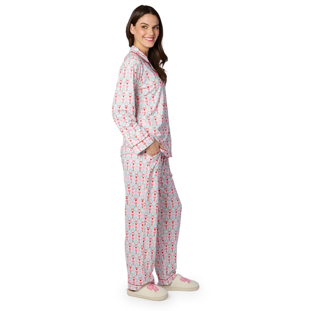 Digital studio photograph of a woman modeling a nutcracker-themed pajama set against a white background, highlighting the red-piped trim, light blue wreath pattern with pink bows, and cozy cream slippers with matching pink ribbon details