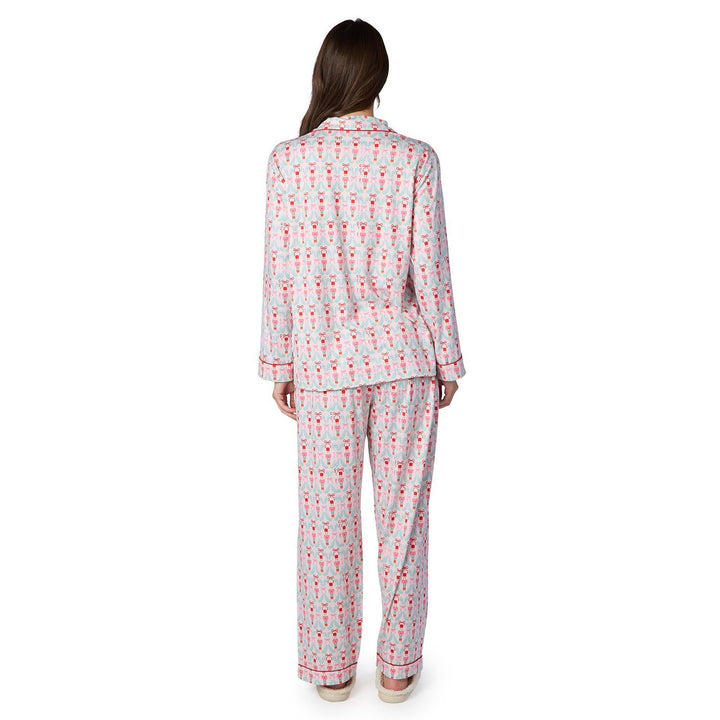 Digital photograph of a woman standing from behind wearing a holiday-themed pink and red nutcracker print pajama set featuring red piping details and plush cream slippers against a plain white background