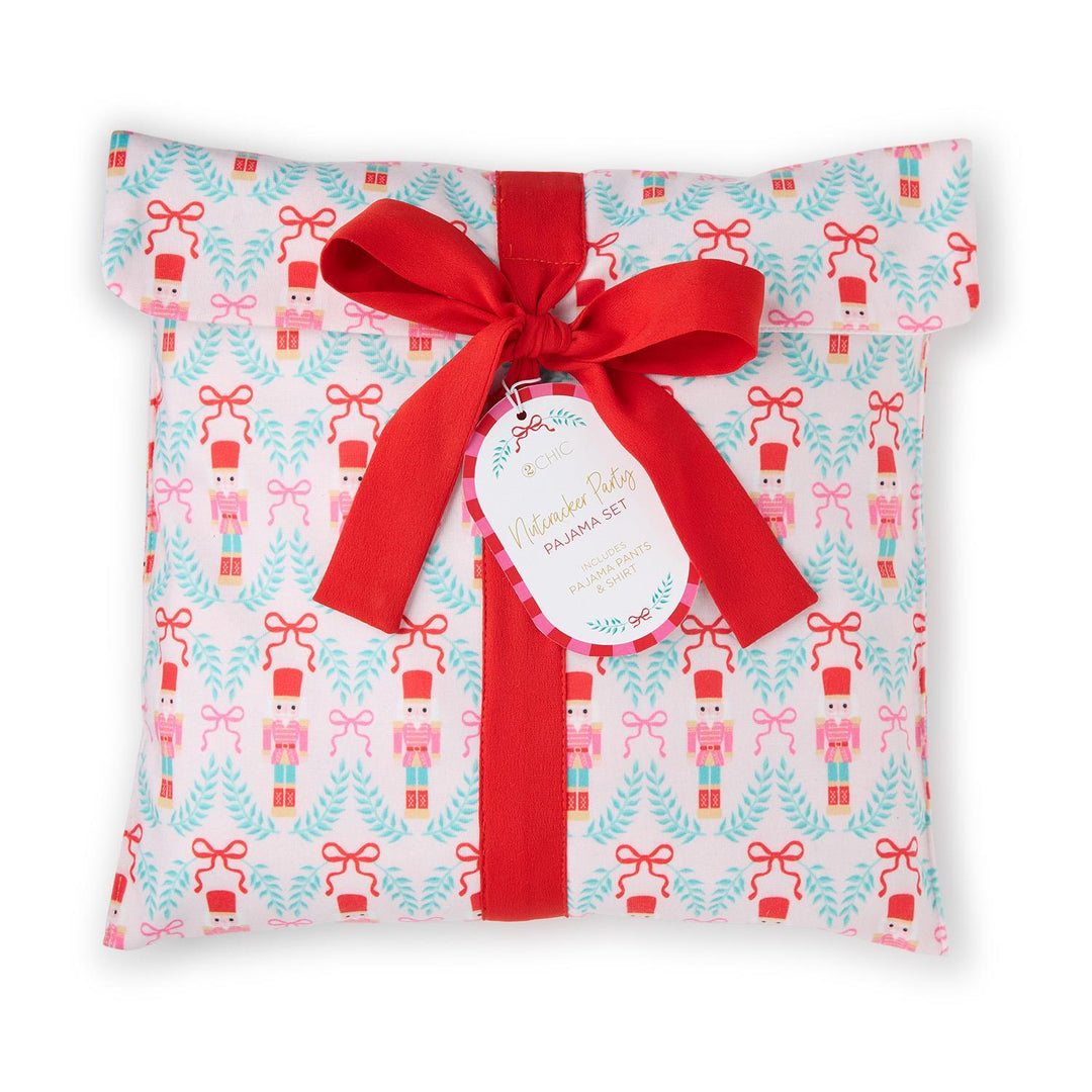 Commercial photograph of a 2CHIC Nutcracker Party pajama set packaged in matching pink fabric with a nutcracker and teal wreath pattern, featuring a large red grosgrain ribbon bow and a circular product tag on a plain white background