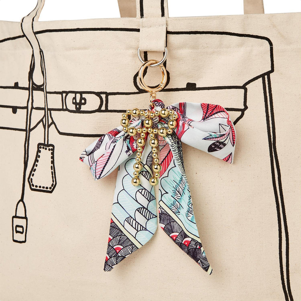 Canvas tote bag featuring a black line art illustration of an iconic handbag silhouette adorned with a gold-beaded bow charm and a colorful patterned ribbon in red and teal