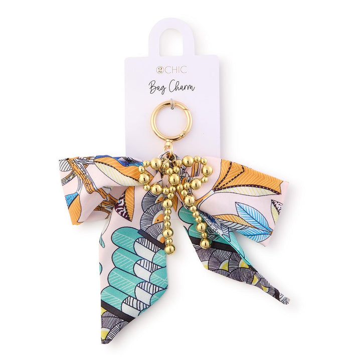 2CHIC fabric and metal bag charm featuring a patterned scarf bow with teal and orange botanical motifs, an intricate gold-beaded bow accent, and a circular gold-toned clasp on a branded white card
