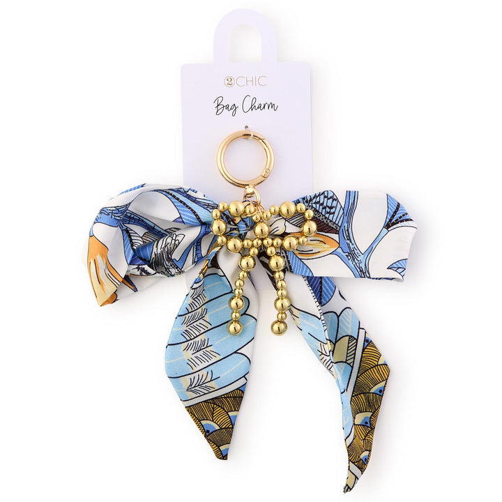 Fabric and metal accessory by 2CHIC featuring a decorative bag charm with a blue and gold botanical print bow and a central gold beaded bow detail on a white display card