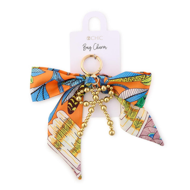 Fabric and metal bag charm by 2CHIC featuring a vibrant orange scarf bow printed with the Eiffel Tower in Paris, leopard pattern accents, and a dangling gold-beaded bow ornament