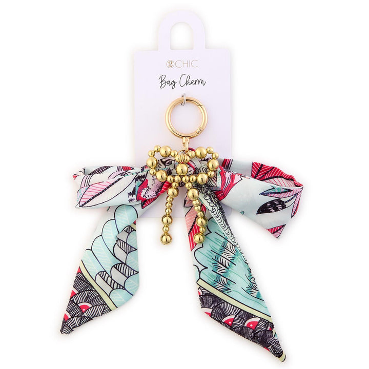 Fabric and metal 2CHIC bag charm on a white display card featuring a light blue bow with red and pink floral patterns, a central gold beaded heart with dangling beads, and a circular gold attachment ring