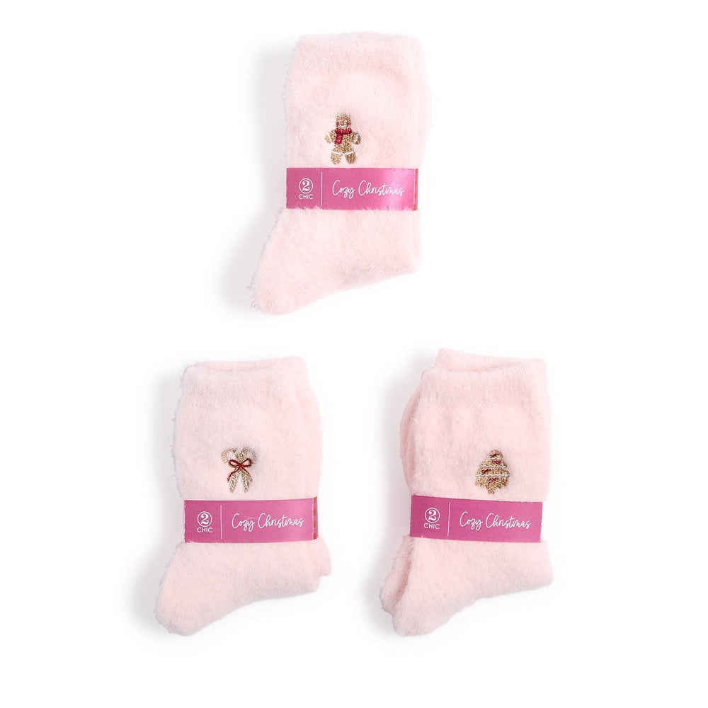 Product photograph by 2 Chic of three pairs of light pink fuzzy socks arranged on a white background, each decorated with gold embroidered holiday icons like a gingerbread man and Christmas tree and wrapped in a magenta paper band labeled Cozy Christmas
