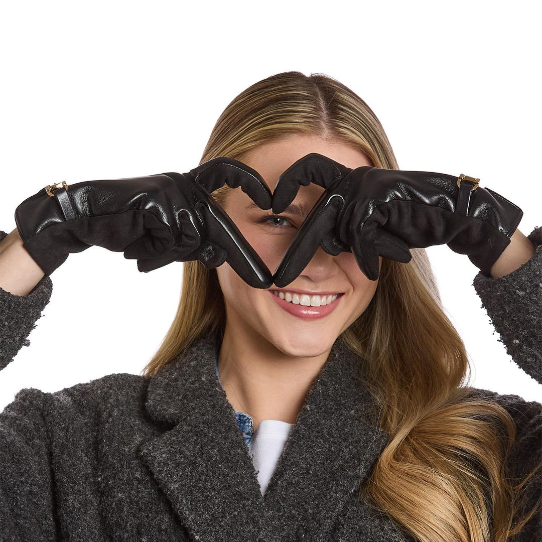 Studio photograph of a smiling blonde woman in a white studio making a heart shape over her face with black leather gloves featuring gold buckle accents and a textured grey wool coat