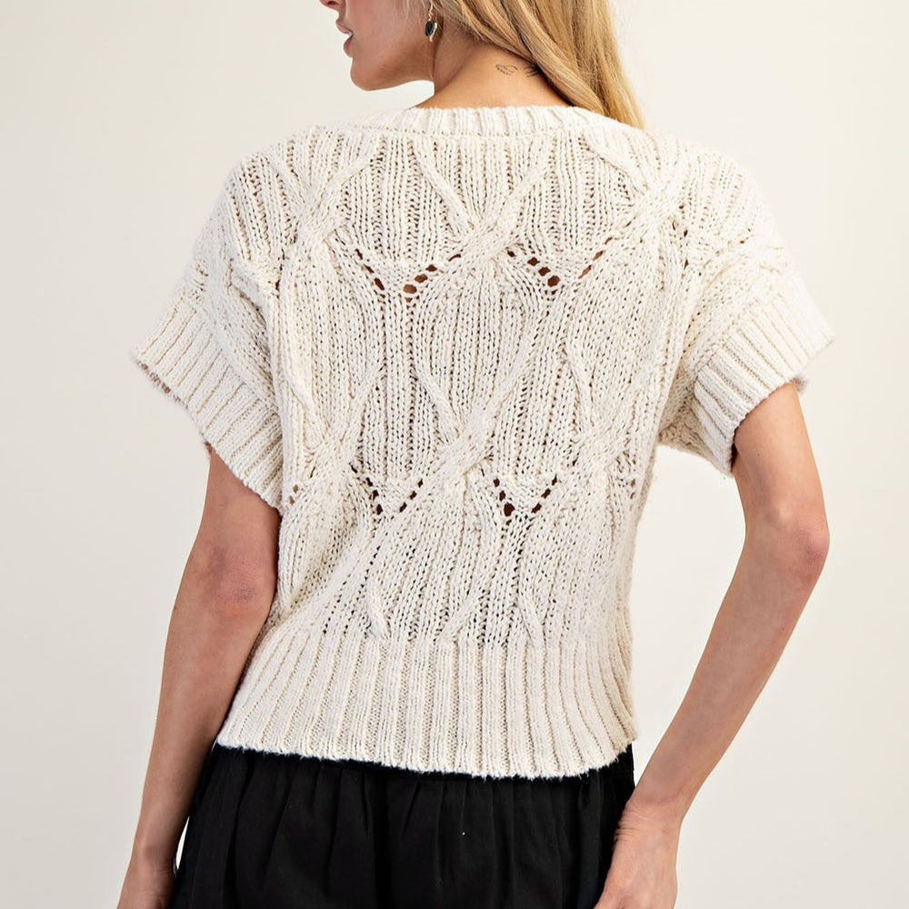 Photograph of a woman wearing a cream cable-knit short-sleeved top in a studio setting, showcasing an intricate diamond-shaped weave, pointelle-style holes, and a wide ribbed hemline