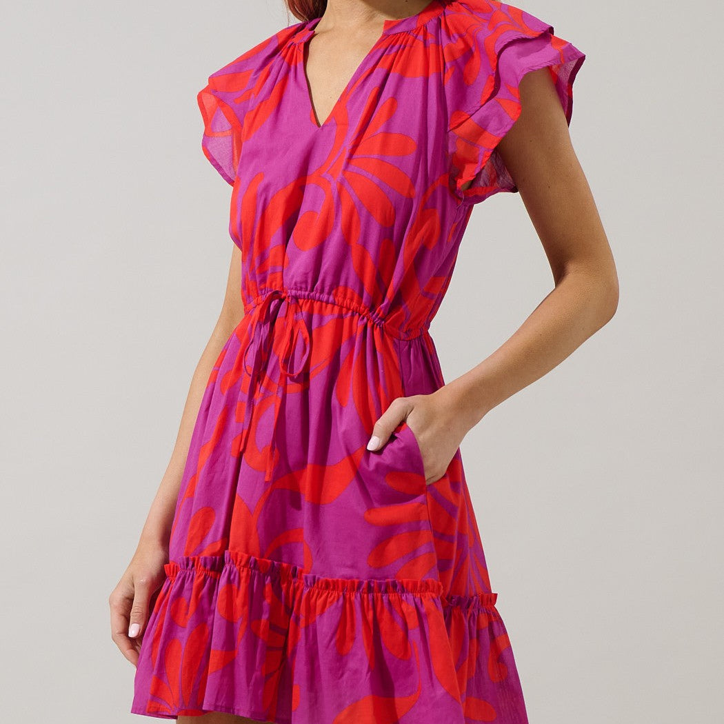 Studio photograph of a model wearing a vibrant pink and red floral patterned dress featuring ruffled flutter sleeves, a drawstring waist, and a tiered hemline against a minimalist grey background